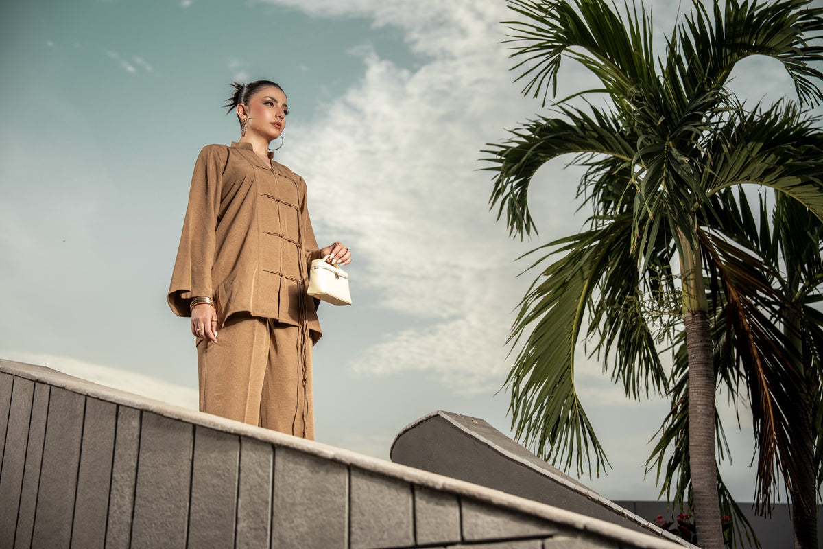 A woman in the Dune sand brown co-ord seen from below against a pale sky, a palm to one side.